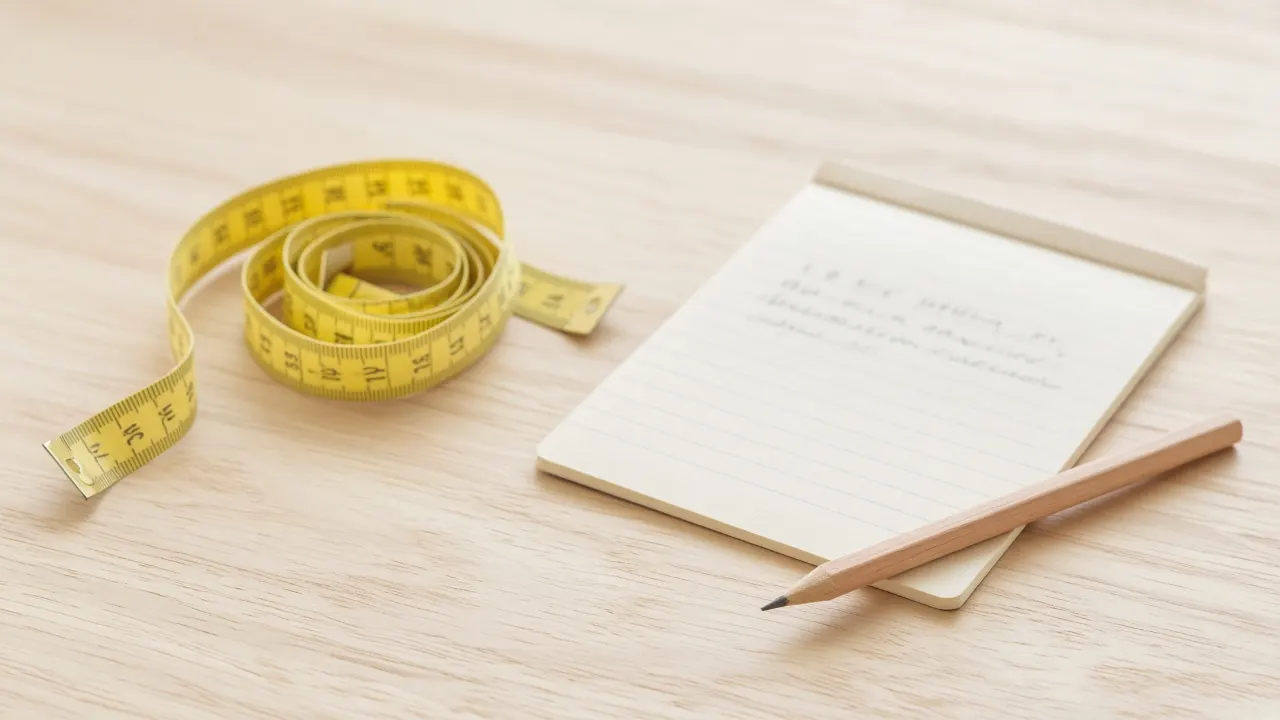 A soft cloth tape measure loosely coiled beside a notepad and pencil on a pale wooden table